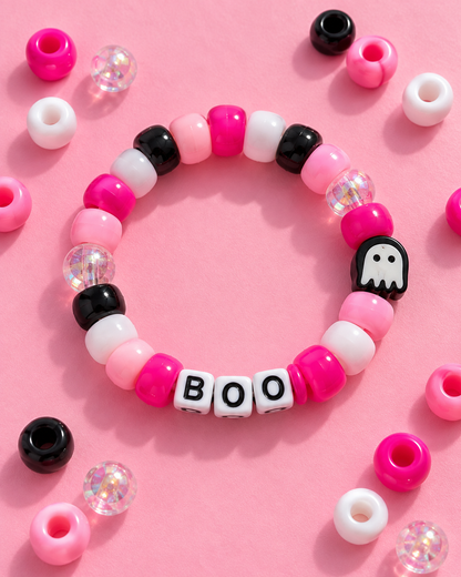 A beaded stretch bracelet arranged in a circle on a pink background, featuring pink, white, and black beads, clear glitter beads, a black ghost charm, and white letter beads spelling BOO.