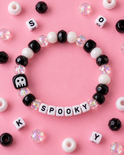A beaded stretch bracelet arranged in a circle on a pink background, featuring black and white beads, iridescent clear beads, and white letter beads spelling 'SPOOKY'. Scattered around are matching loose beads and letter cubes.