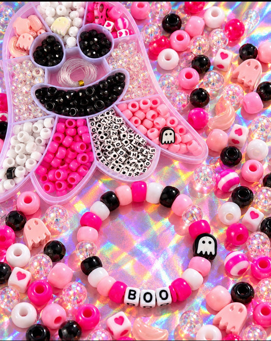 A Halloween-themed bead craft kit and matching bracelets arranged on a holographic background. The kit includes a pink monster-shaped organizer filled with black, white, and pink beads, plus several beaded bracelets featuring pink, white, and black beads, ghost charms, and a white letter bead spelling 'BOO'.