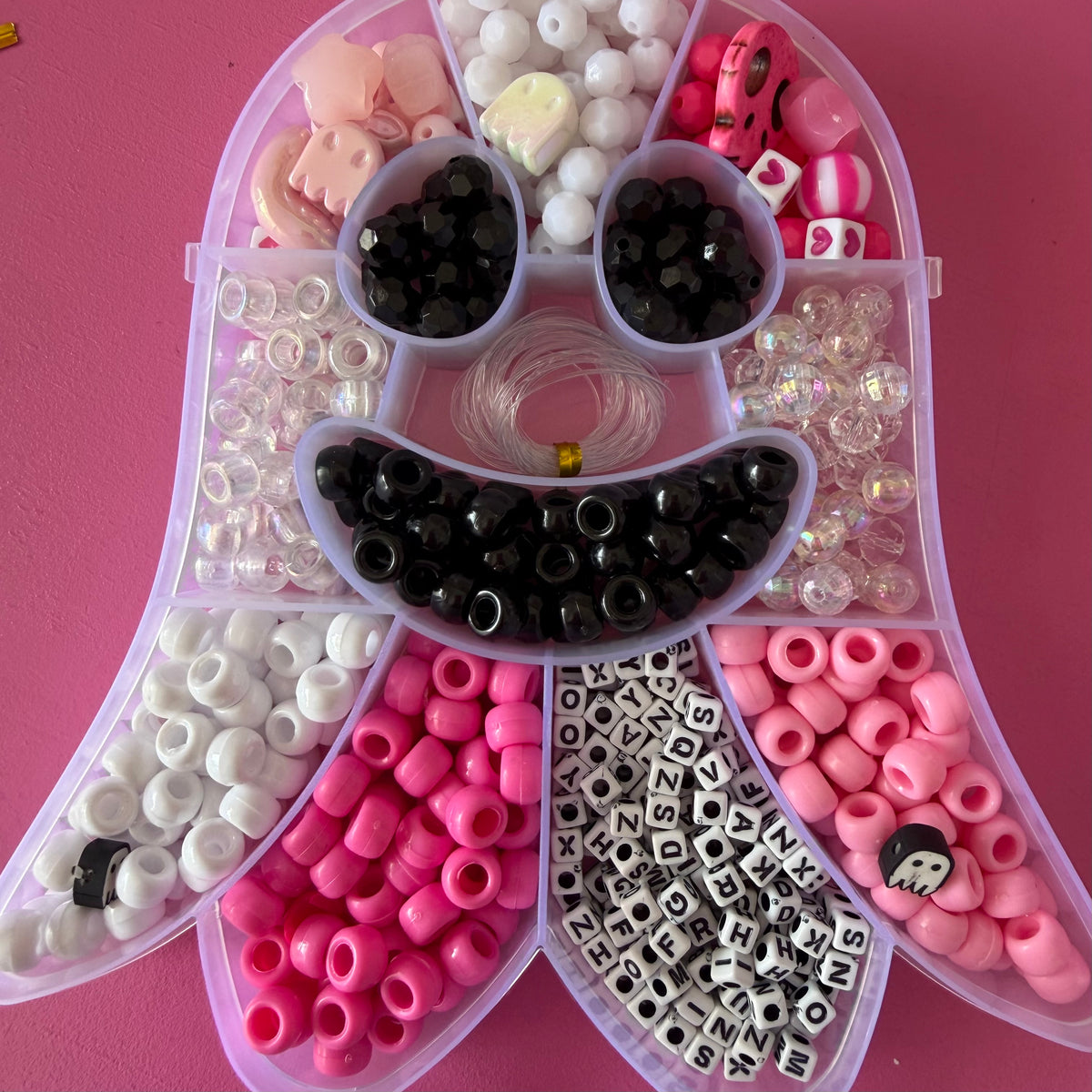 A clear plastic organizer tray filled with assorted craft beads and letter tiles in pink, white, black, and iridescent finishes, arranged on a solid pink background.