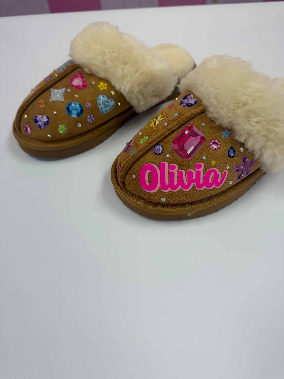 Pair of personalized chestnut faux-fur slippers covered in colorful gem designs, one with Olivia in pink