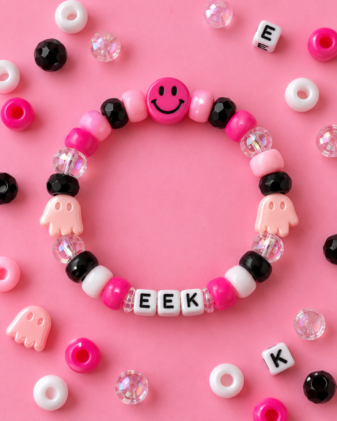 A beaded bracelet arranged in a circle on a pink background, featuring pink, black, and white beads, a smiling face charm, ghost charms, and letter beads spelling 'EEK'.